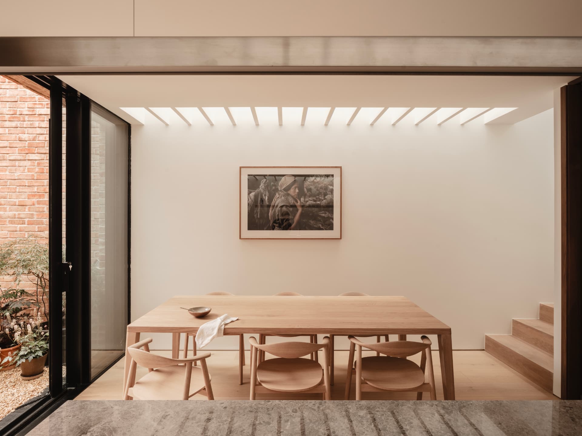 A bright dining area in an 1880s Dublin home, featuring a custom oak dining table and chairs, a framed photograph, a linear skylight, and sliding glass doors looking out to a courtyard.